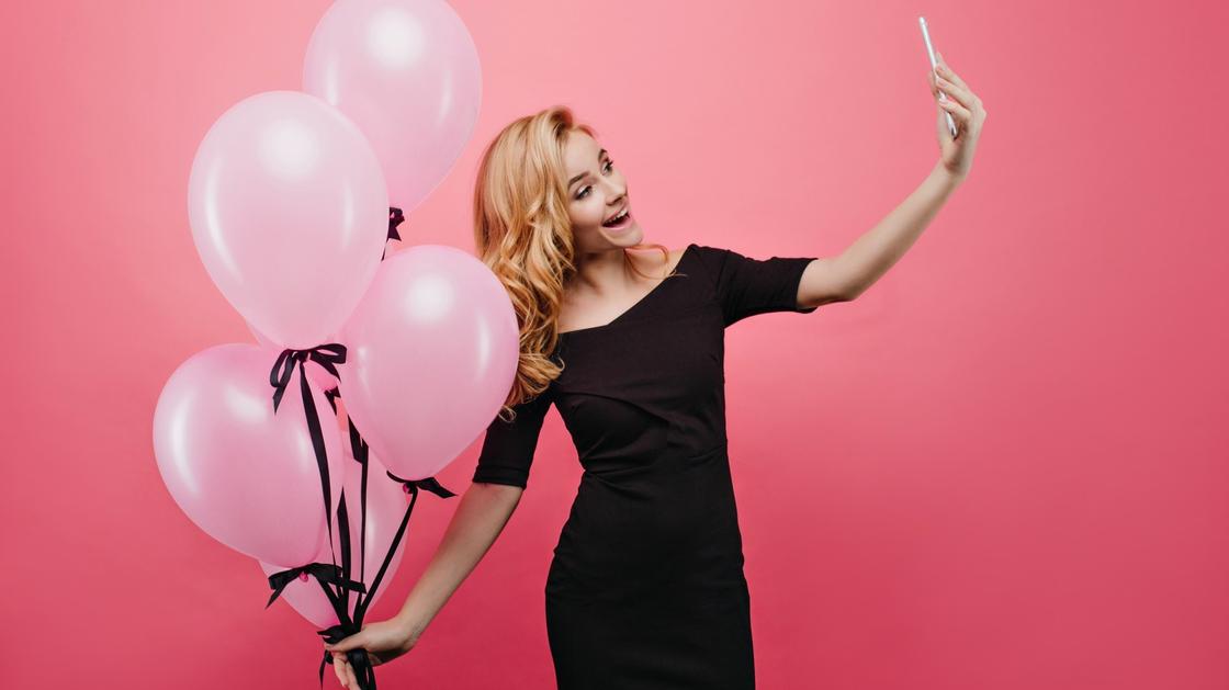 Девушка в черном платье улыбается и сомтрит в экраг телефона. В руке она держит связанные вместе розовые воздушные шарики