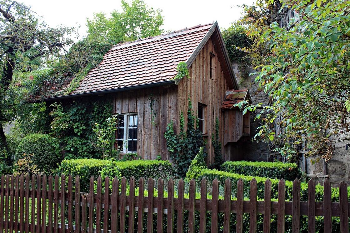 Старый деревянный дом ав окружении зеленых кустов и деревьев. Перед домомо установлен забор из деревянного штакетника