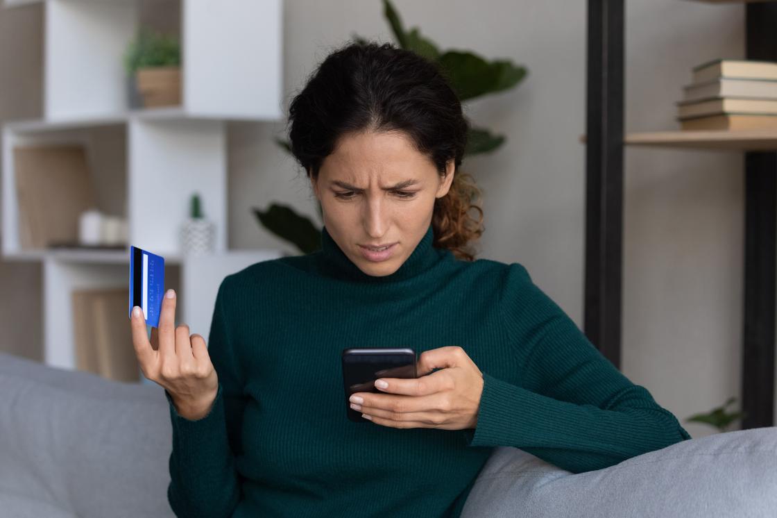 Девушка смотрит в экран телефона, держа в руках банковскую карту