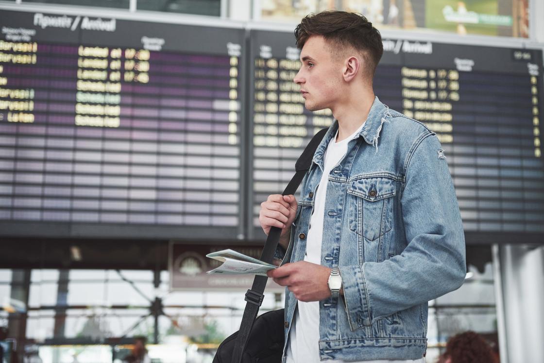 Мужчина с серьезным взглядом в джинсовой куртке на фоне табло прибытия аэропорта