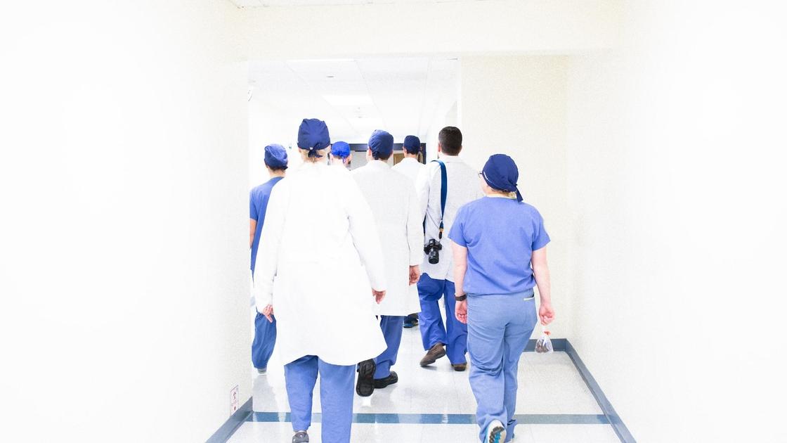 Несколько медиков в халатах и спецодежде уходят по коридору к открытой двери. У одного из них висит фотоаппарат через плечо