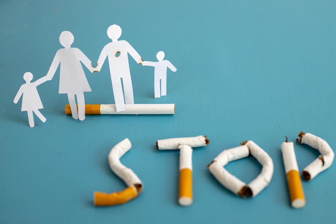 Прожженные бумажные фигурки семьи стоят рядом с надписью "STOP" из сигарет