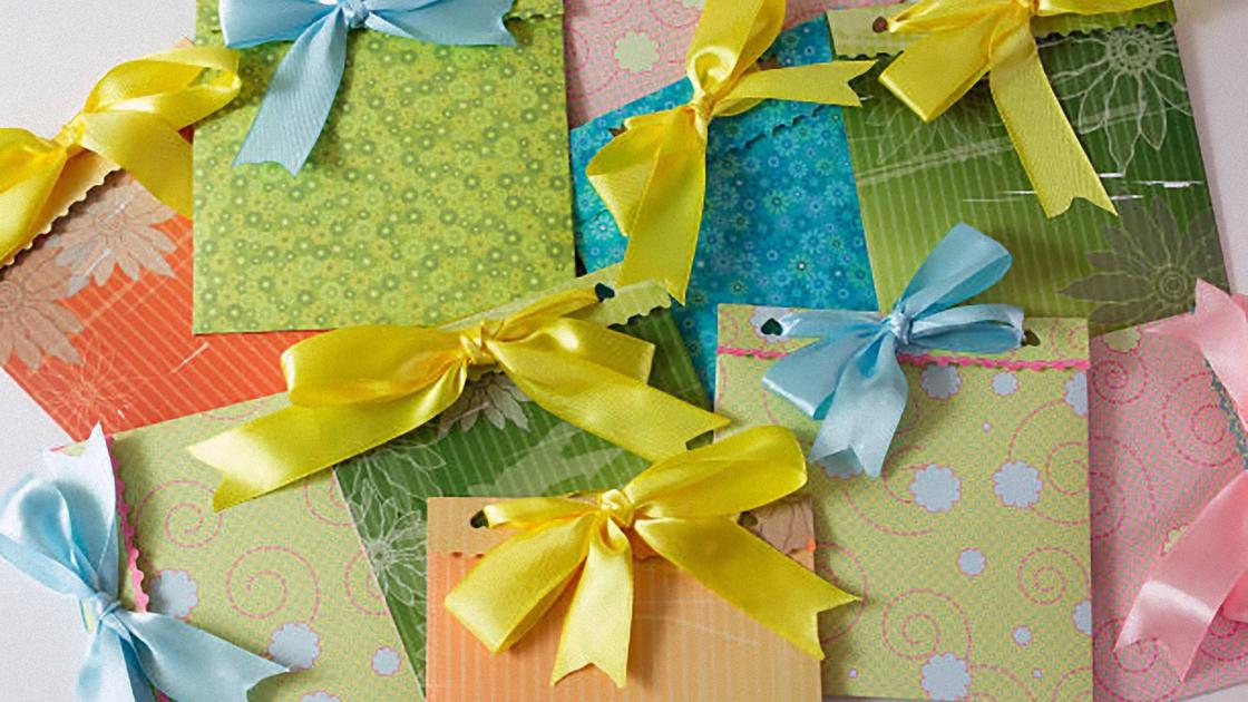На столе лажат конверты с разноцветной скра-бумаги, украшенные сверху желтыми и голубыми бантиками