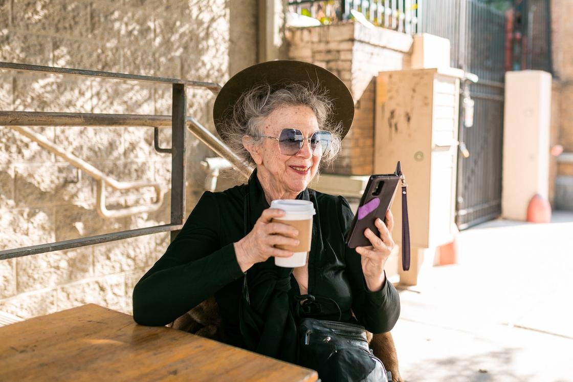 Женщина за 60 сидит в чашкой кофе и телефоном