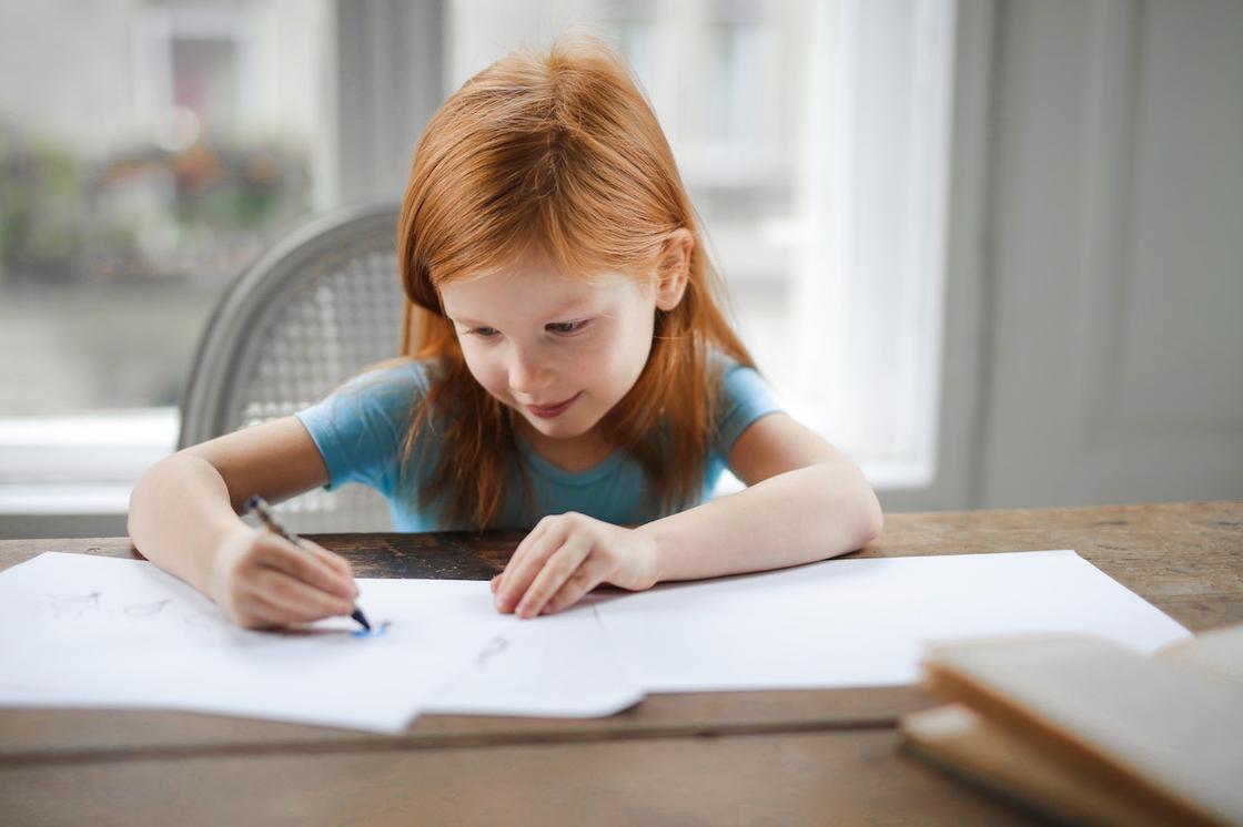 Рыжая девочка рисует в тетради, сидя за столом
