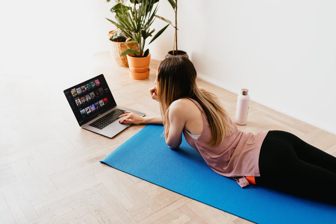 Девушка в спортивной одежде лежит на коврике для йоги и смотрит на экран ноутбука