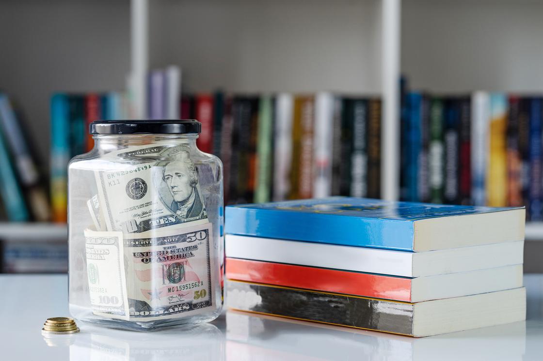 Стеклянная банка с долларами стоит возле стопки книг