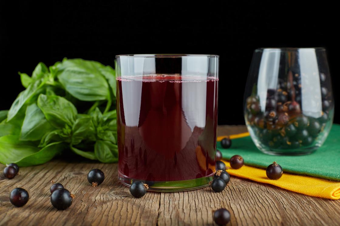 На столе стакан с красным напитком и ягоды черной смородины