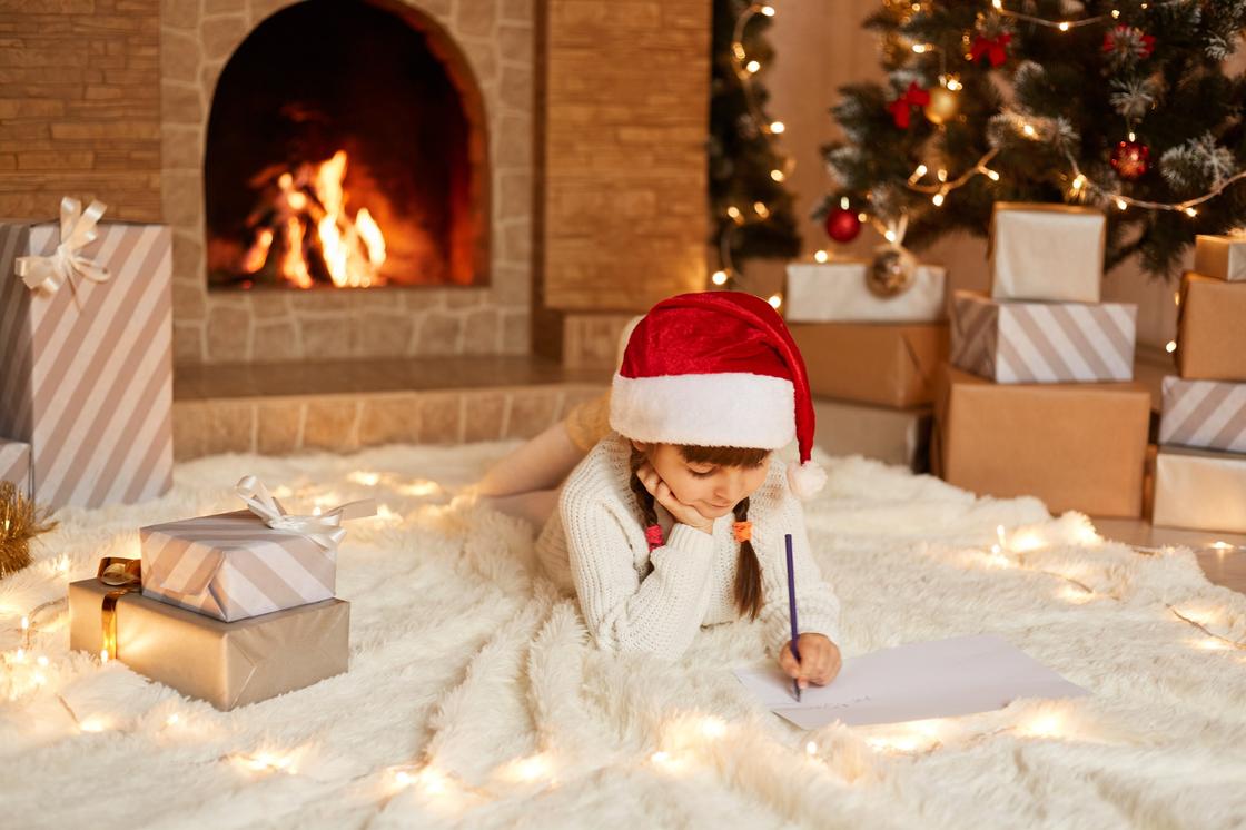 Девочка лежит на мягком пледе у елки и коробок с подарками и пишет письмо Деду Морозу