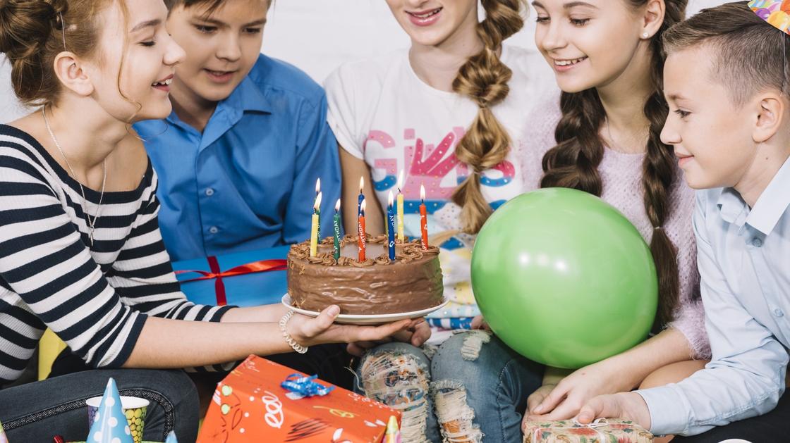 Девочка держит в руках торт со свечами. Вокргу сидят дети с воздушными шариками и в колпачках и улыбаются