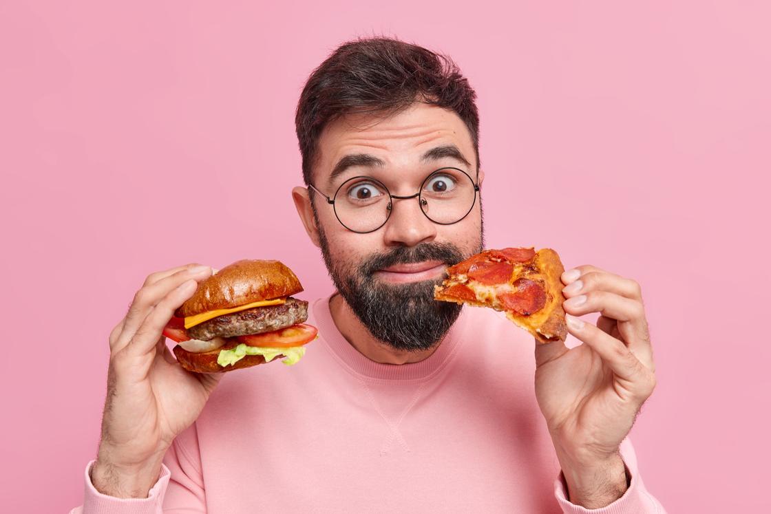 Бородатый мужчина в очках держит бургер и кусочек пиццы