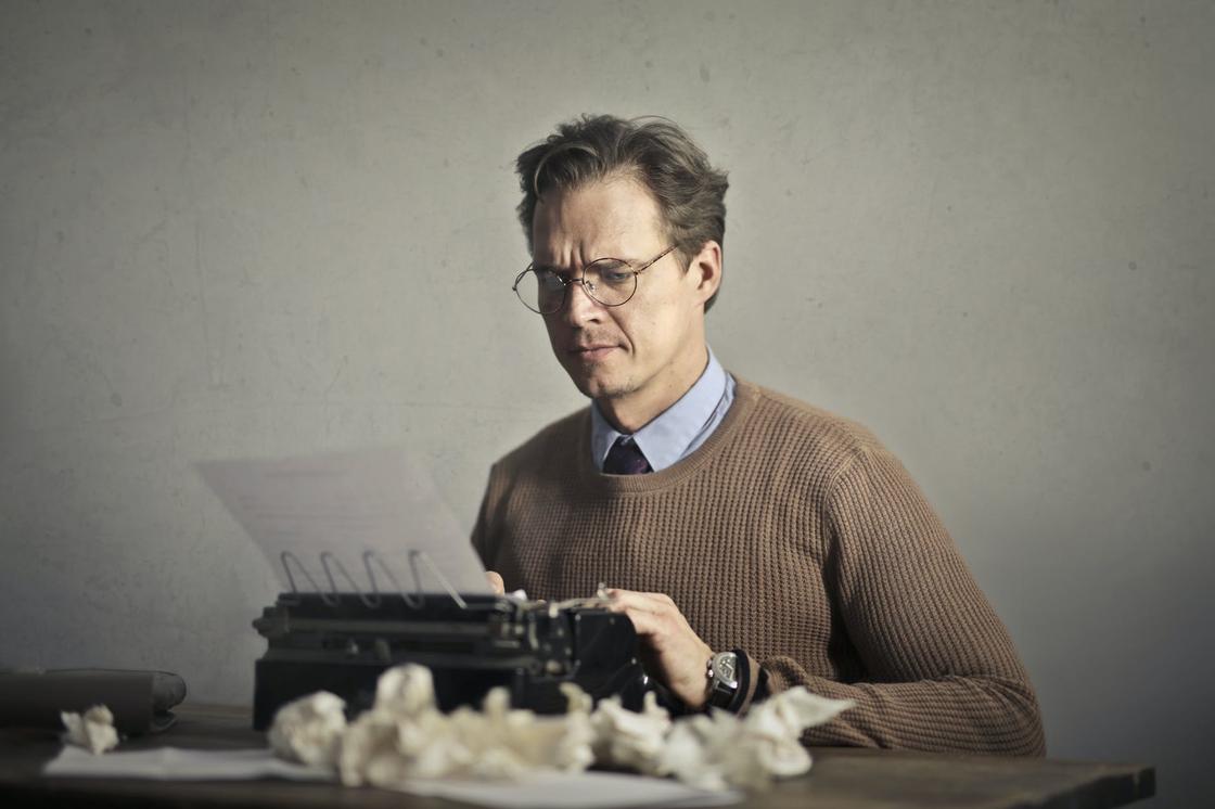 Мужчина в очках и коричневом свитере печатает на печатной машинке