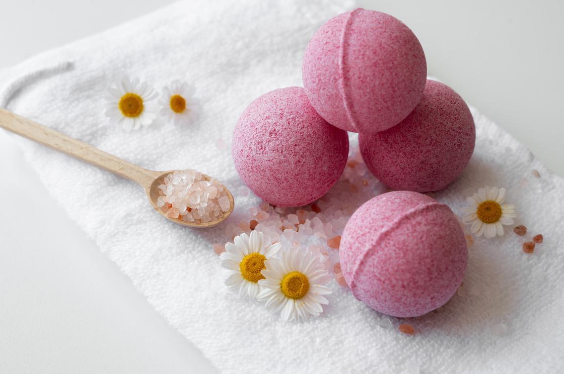 Розовые бомбочки для ванны лежат на полотенце. Рядом рассыпаны цветы ромашки и розовая соль в деревянной ложке