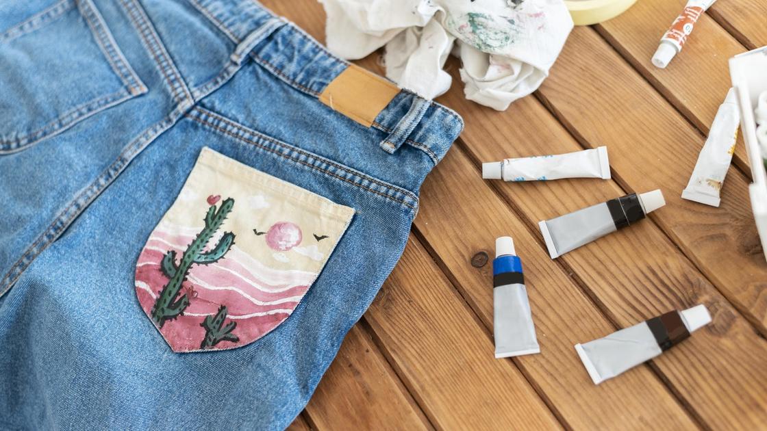 На заднем кармане джинсов сделана нашивка с летним пейзажем. Рядом лежат тюбики краски и тряпки