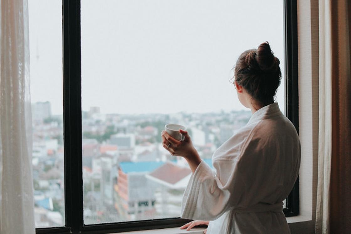 Девушка в белом халате с чашкой в руке смотрит в окно