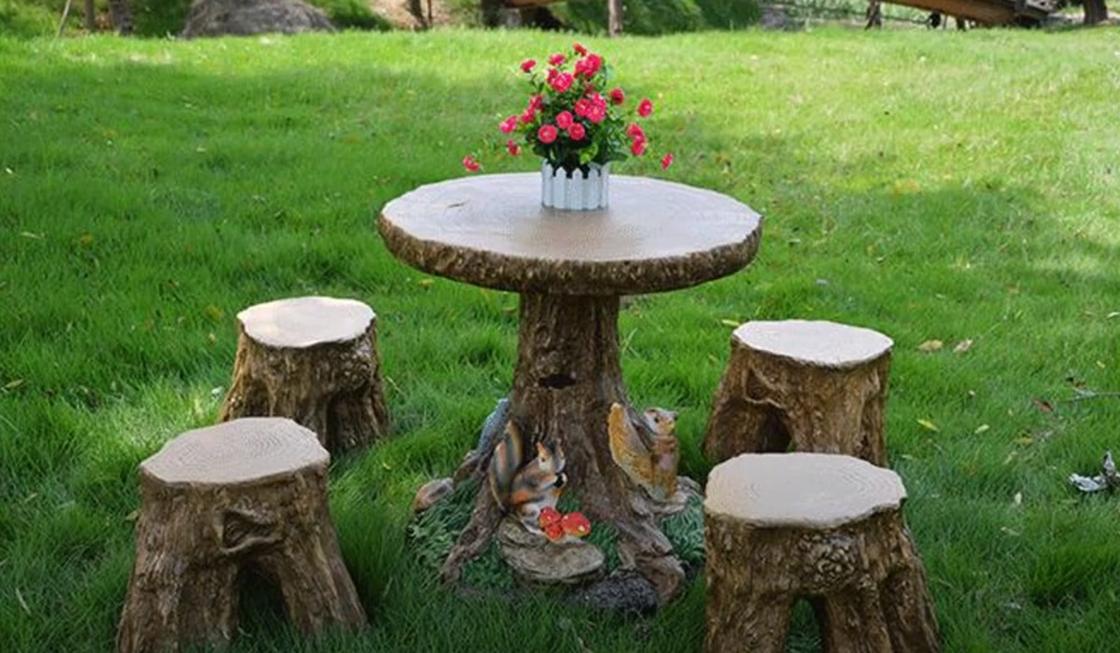 Столик и четыре стула из пеньков стоят на зеленой лужайке. На столе стоит ваза с цветами