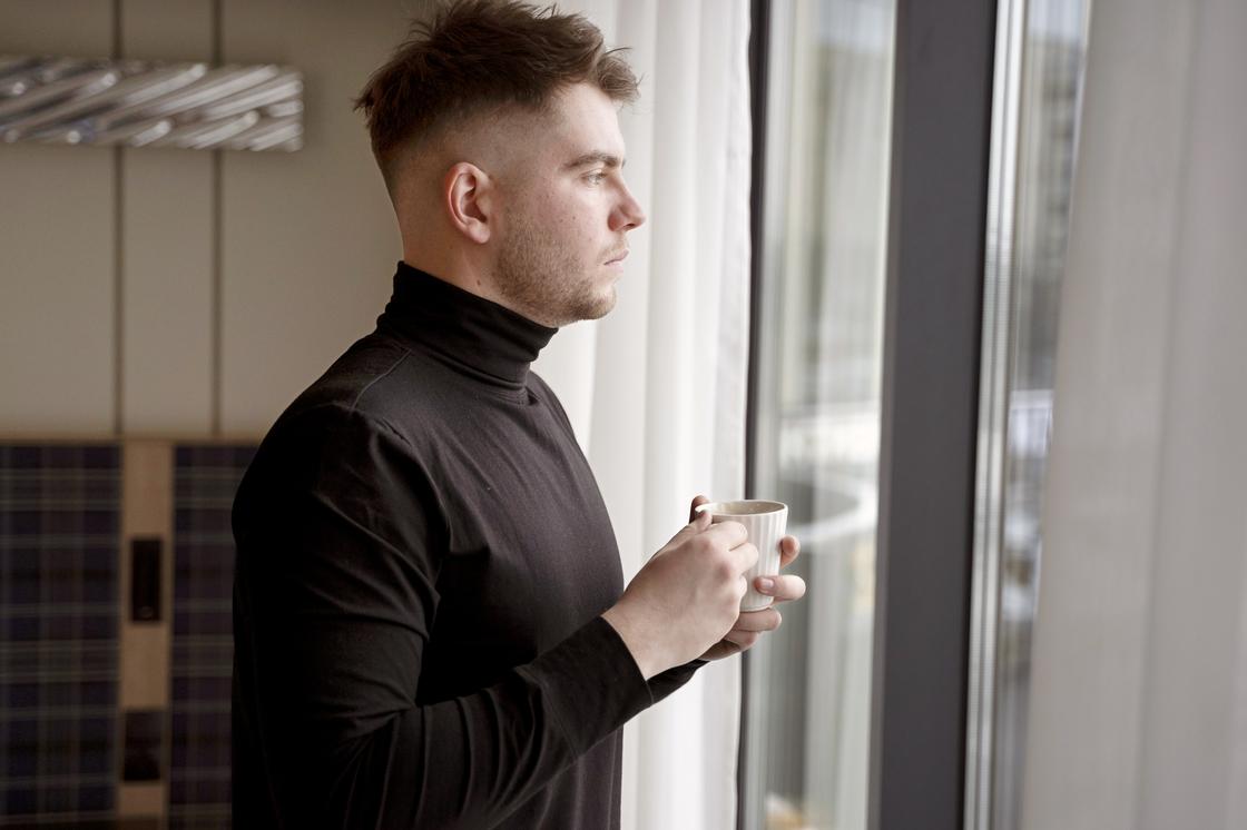 Задумчивый мужчина стоит перед окном с чашкой кофе
