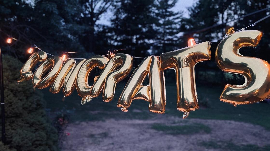 Позравительные воздушные шары в виде надписи «Congrats» развешены на светящейся гирлянде в саду