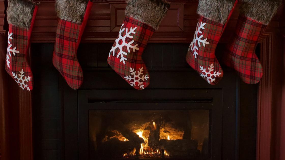 Новогодние носки висят над камином, где горит огонь. Носки сделаны из клетчатой красной ткани, украшены мехом и белыми снежинками