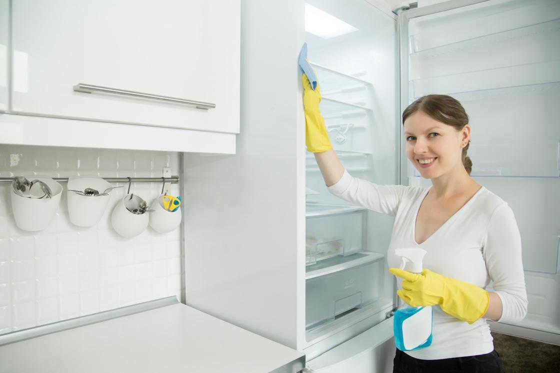 Девушка в желтых резиновых перчатках протирает салфеткой холодильник внутри. В другой руке она держит пульверизатор с жидкостью