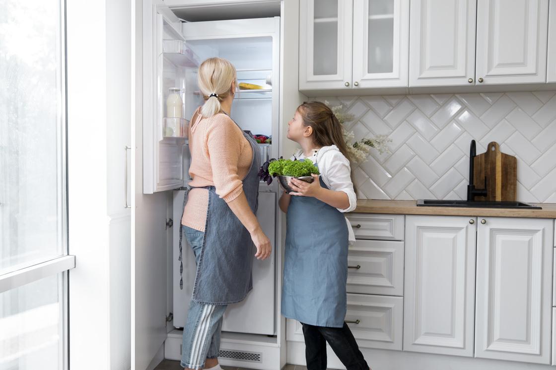 Женщина и девочка в фартуках заглядывают в холодильник на кухне. В руках девочка держит миску с зеленью