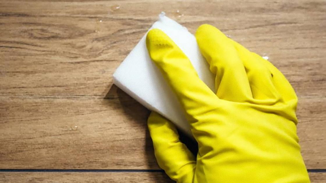 Рукой в желтой резиновой перчатке держат меламиновую губку и вытирают поверхность