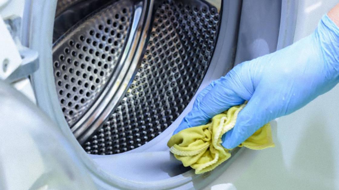 В открытом люке стиральной машины видно барабан. Его протирают рукой в перчатке с помощью желтой салфетки