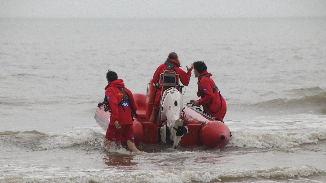 Три спасателя в красных спецкостюмах выводят моторную лодку в море