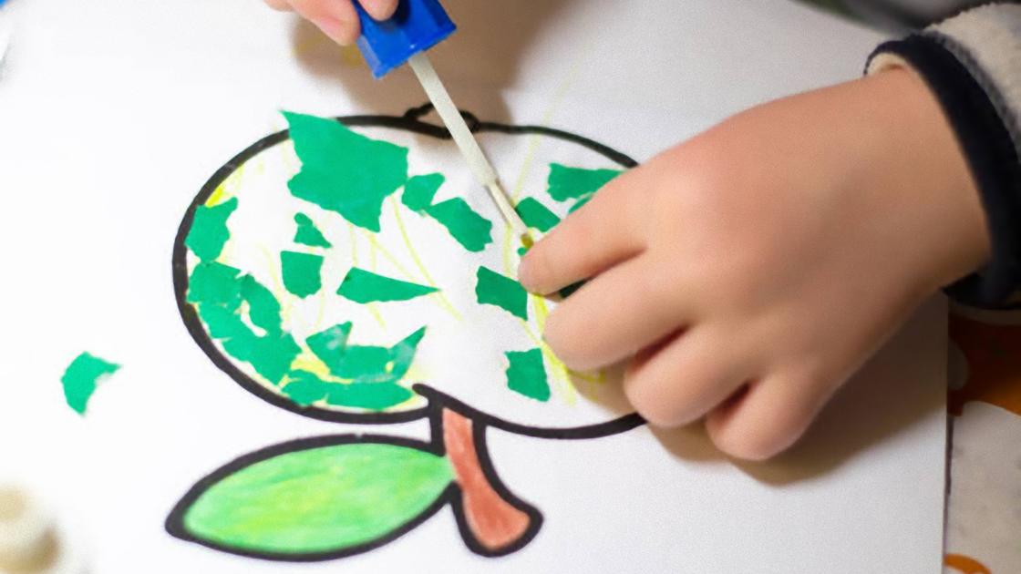 Ребенок приклеивает кисточкой к нарисованному на бумаге яблоку кусочки рваной зеленой бумаги