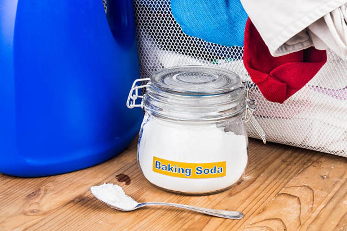 Сода в стеклянной банке с крышкой и в ложке находится рядом с корзиной с бельем и синей пластиковой емкостью