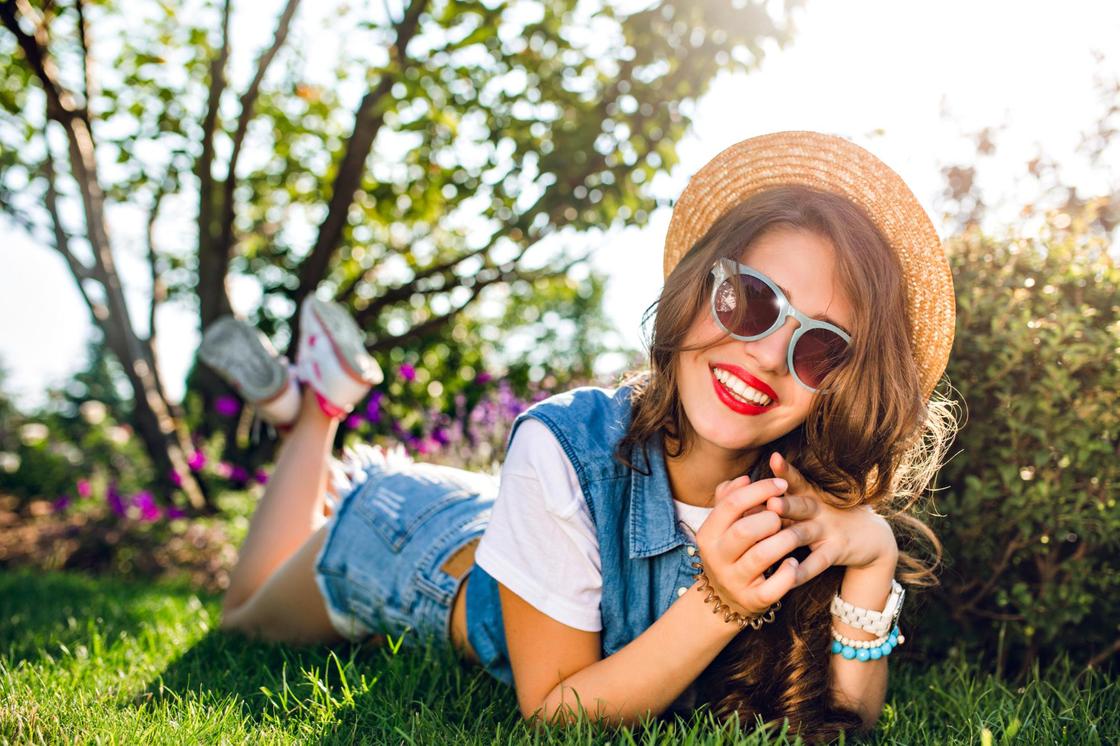 Красивая девушка в очках, шляпе и джинсовой одежде лежит на траве