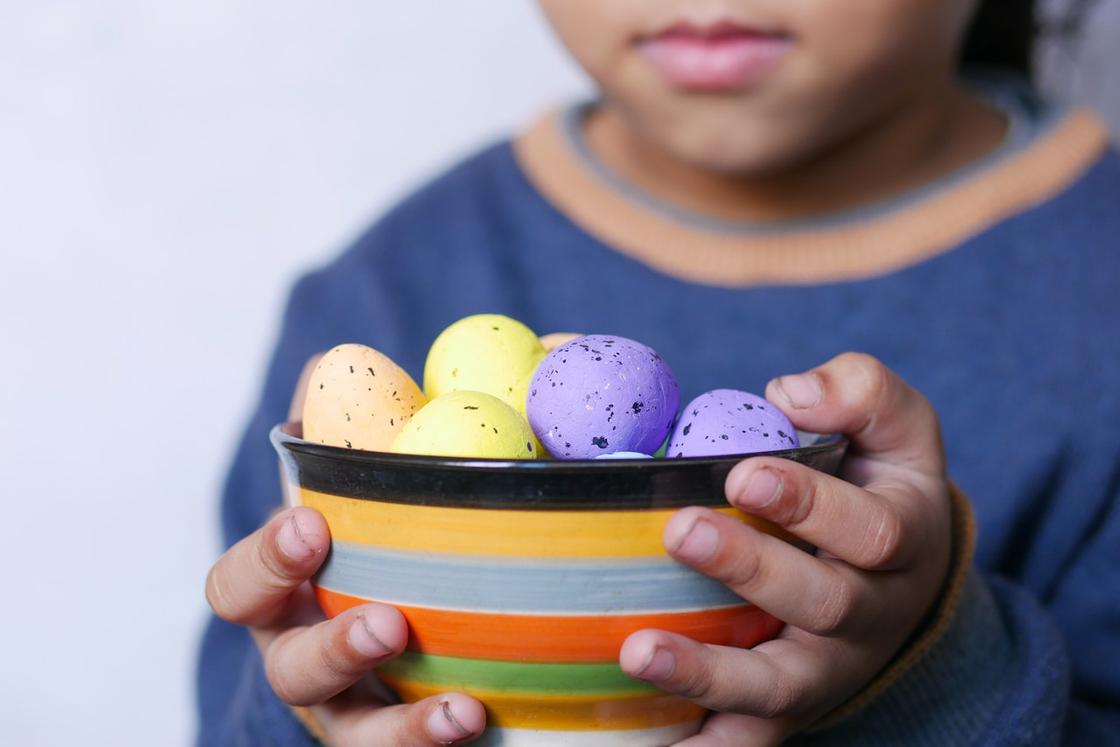 Ребенок держит в руках тарелку с разноцветными яйцами