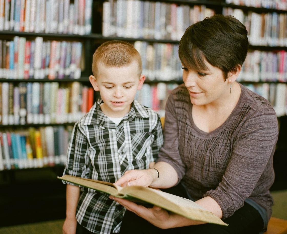 Женщина с короткой стрижкой сидит в библиотеке и читает книгу мальчику. Мальчик одет в клетчатую рубашку