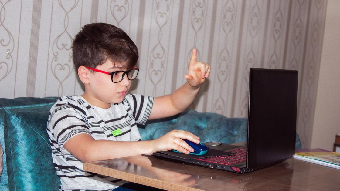 Мальчик в очках и полосатой футболке сидит за столом с открытм ноутбуком. Указательный палец левой руки он поднял вверх, а правой водит мышку на открытом ноутбуке
