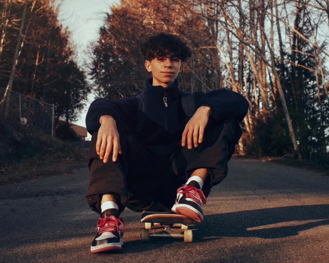 Подросток сидит на земле и придерживает ногой скейтборд