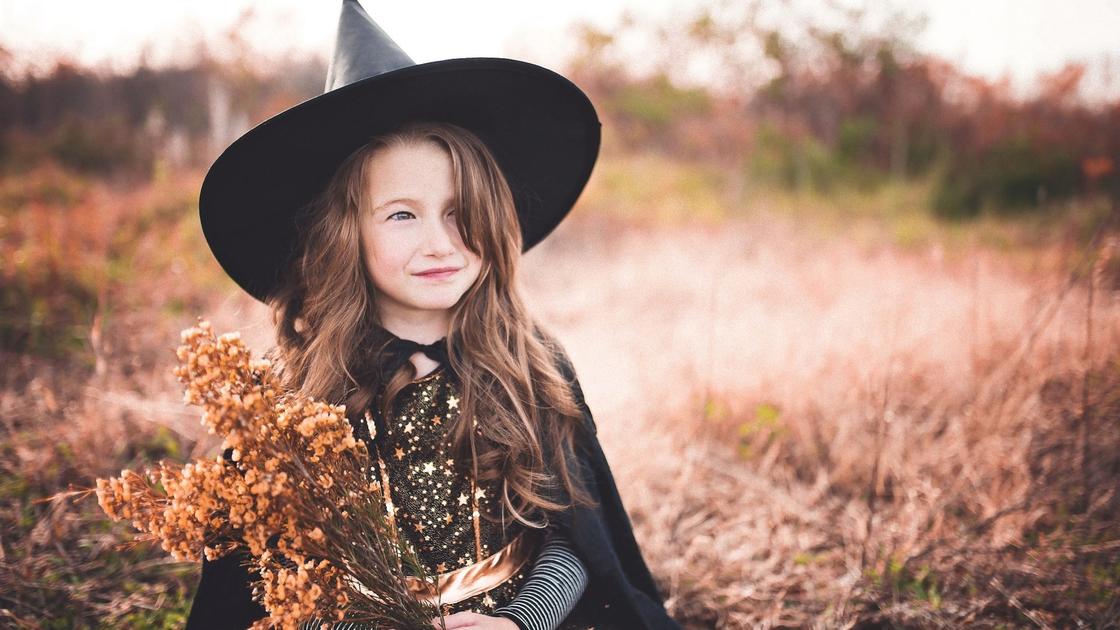 Девочка с длинными волосами в черной шляпе и накидке с букетом полевых цветов сидит на лужайке
