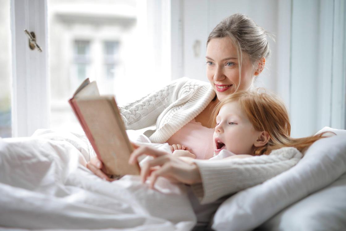 Мама читает дочке книгу, сидя на диване и укутавшись в плет