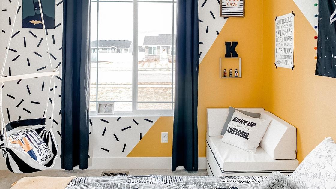 Стены в комнате сделаны в желтом цвете и черных линиях на белом фоне. На окнах висят синие шторы, на стенах висят тематические картины, календарь, возле кровати стоит удобный мягкий уголок, висит качель