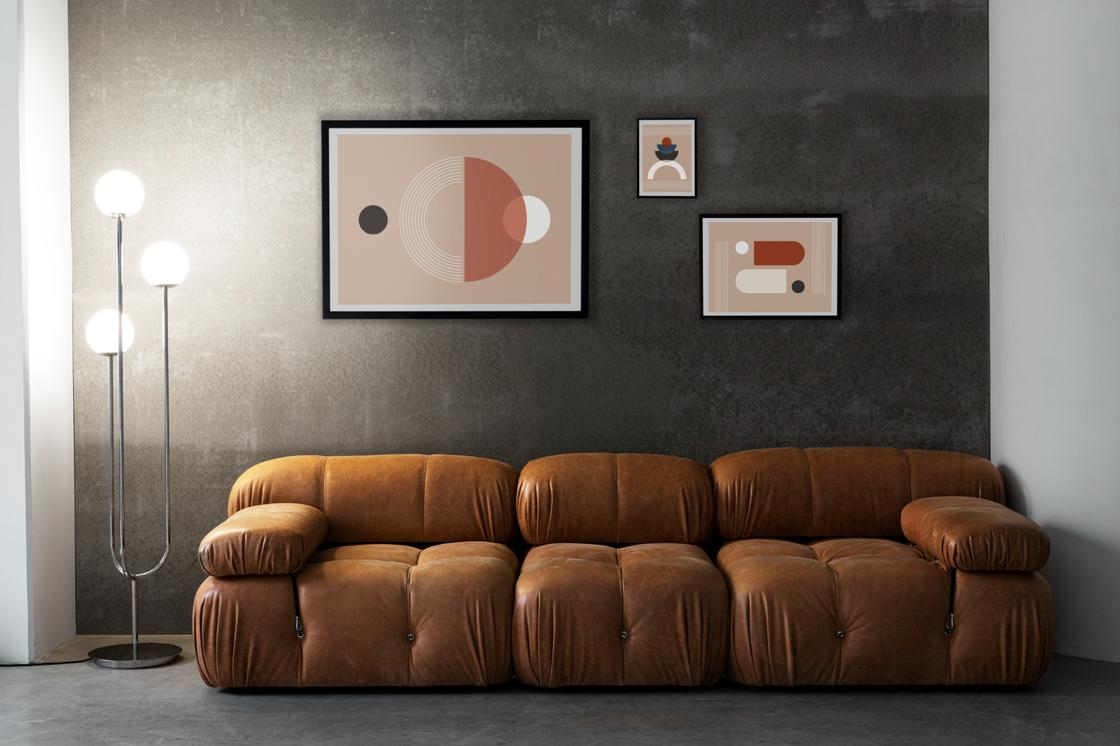 В комнате кожаный коричневый диван, темная стена, торшер и картины