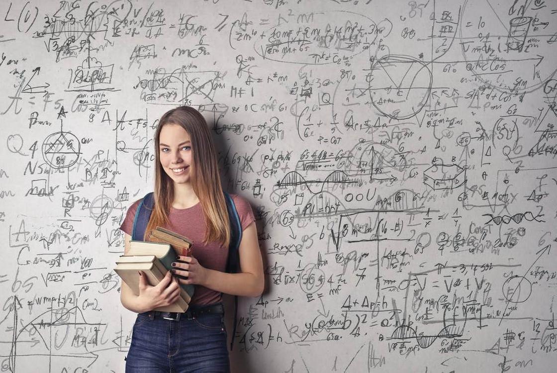 Девушка с книгами стоит возле доски, исписанной математическими формулами