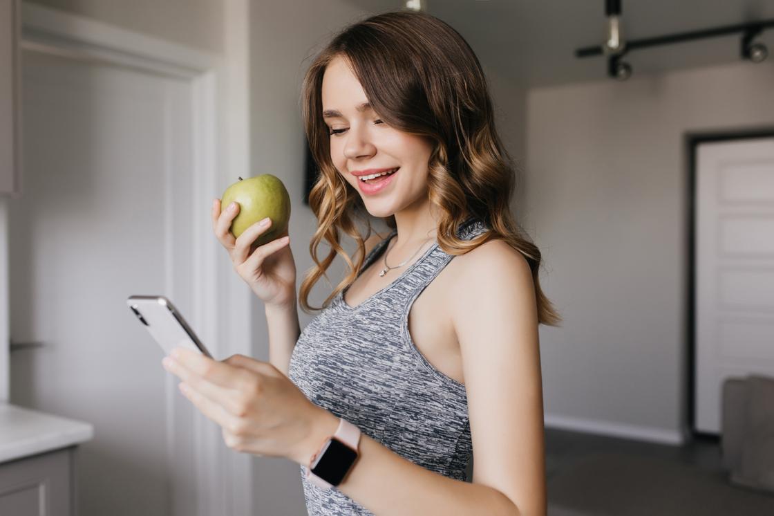 Девушка улыбается, держит в руке яблоко и смотрит в смартфон