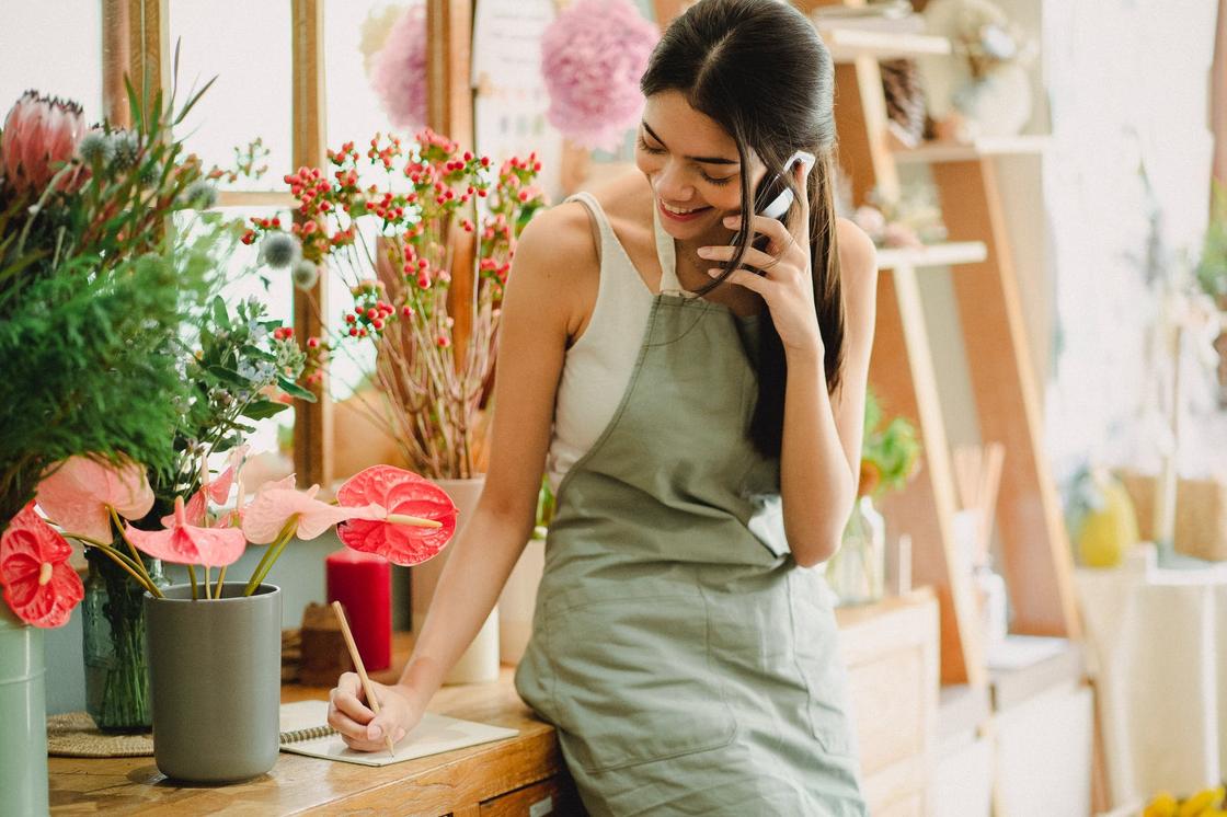 Девушка в фартуке в цветочном магазине разговаривает по телефону и записывает заказ, сидя на столе