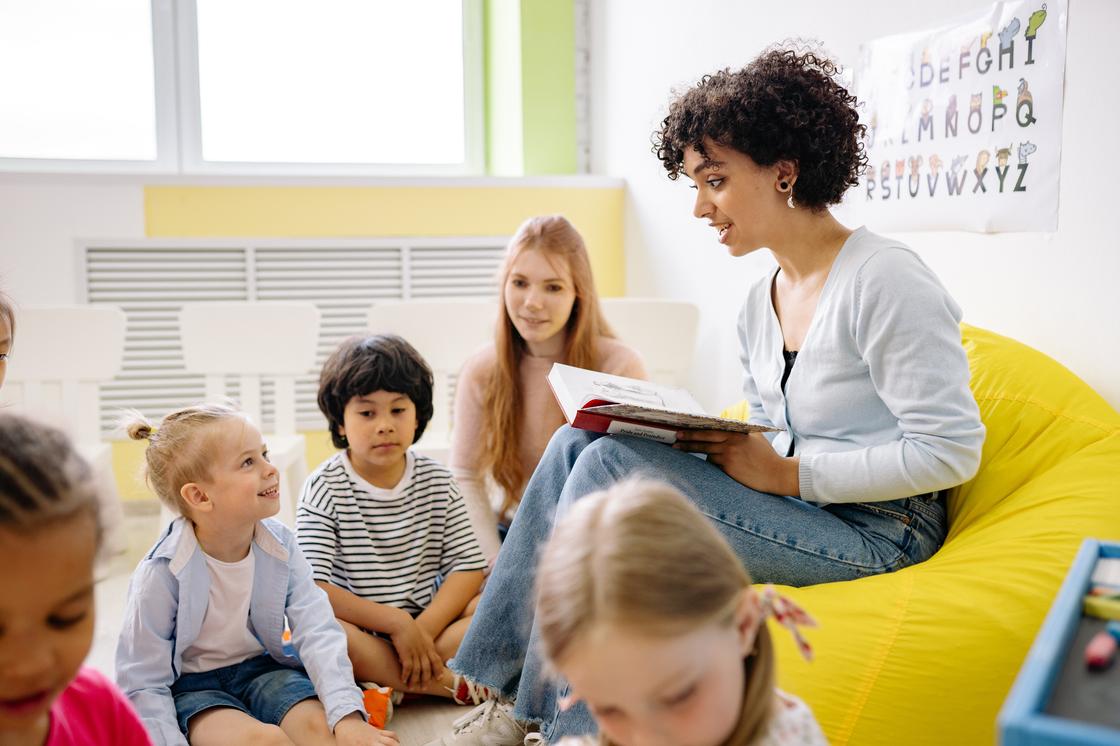 Дети слушают учительницу, сидящую с книгой на коленях
