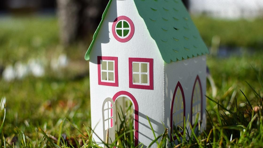 Бумажный домик с зеленой крышей, красными наличниками на окнах и открытой дверью стоит в траве