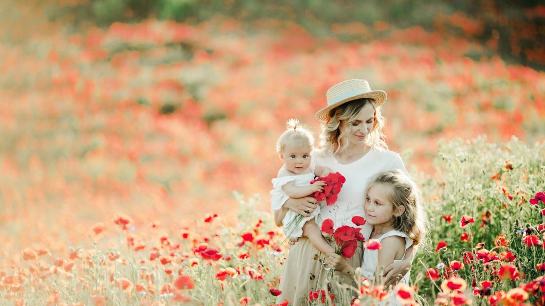 Женщина в шляпе держит на руках маленькую девочку, а вторую прижимает к себе. Они стоят на лугу с цветущими красными маками
