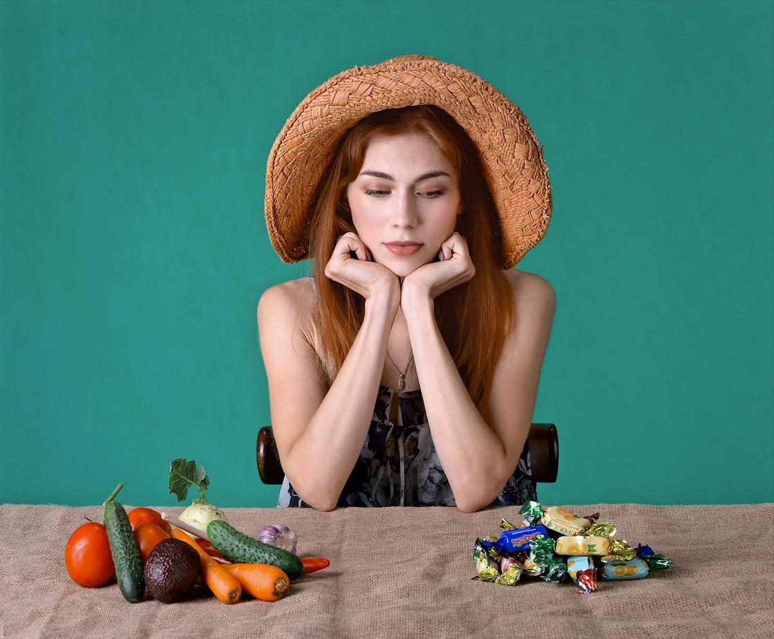 Девушка сидит за столом, на котором лежат морковь, огурцы, помидоры, авокадо, чеснок и конфеты