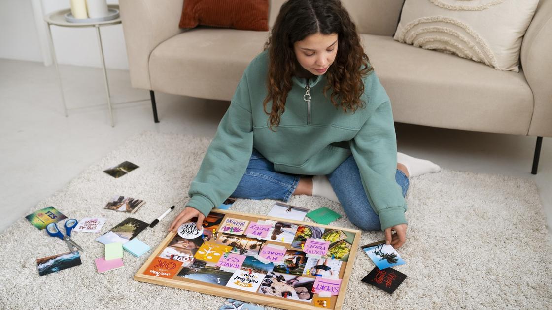 Девушка в толстовке сидит на полу и раскладывает фотографии и картинки с надписями на доске