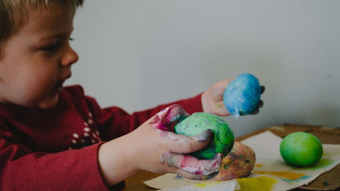 Мальчик держит в правой руке шар зеленого пластилина, а в левой руке у него синий пластилин. Разноцветные шары пластилина лежат перед ним на листке бумаги