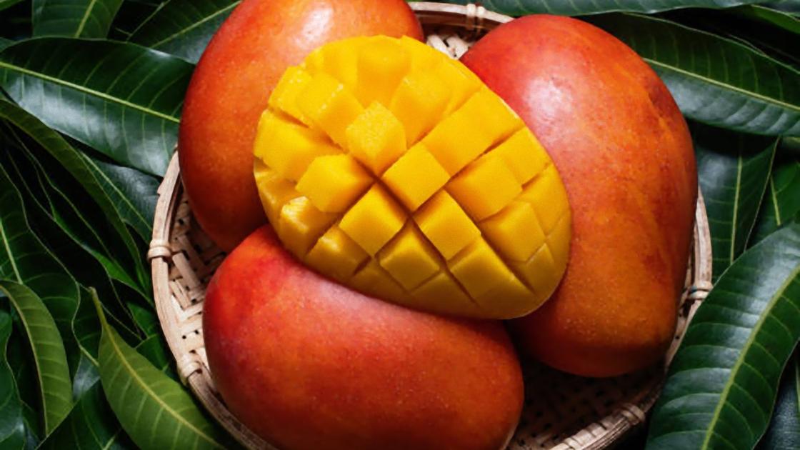 В плетеной вазе лежат три целых плода манго и сверху нарезка из половинки манго