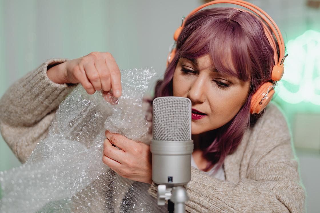 Девушка с яркими волосами в наушниках лопает пузырьки защитного пакета возле микрофона